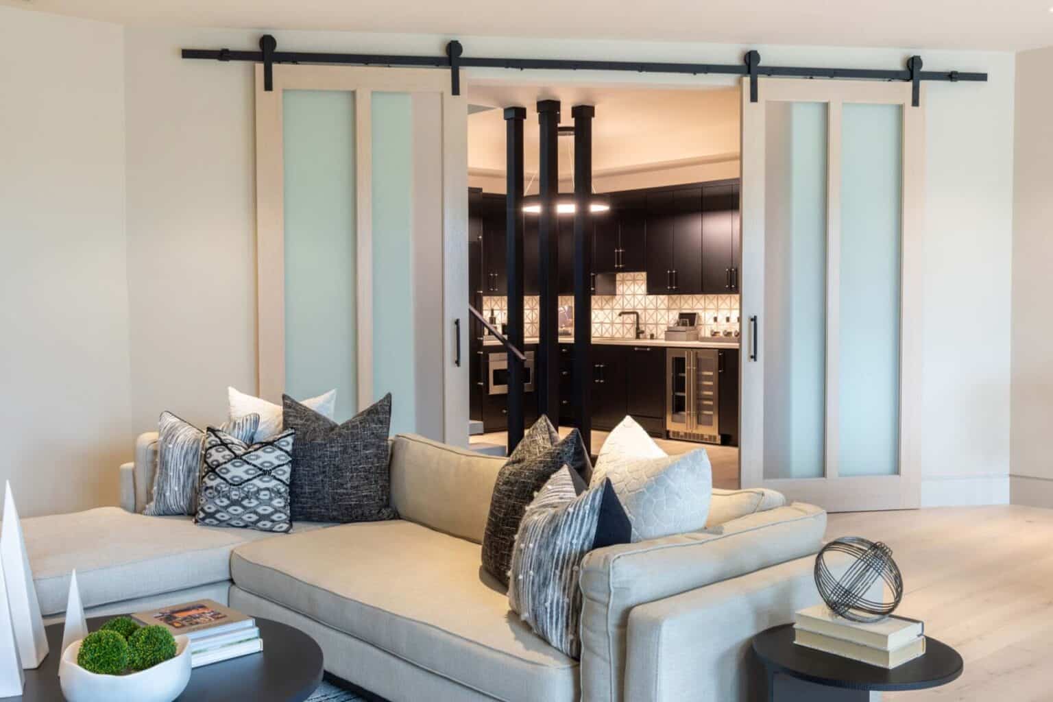 Modern living room with a beige sectional sofa and decorative pillows, adjacent to a kitchen separated by sliding barn doors with frosted glass panels, showcasing expert custom finish carpentry Bellevue homeowners admire.
