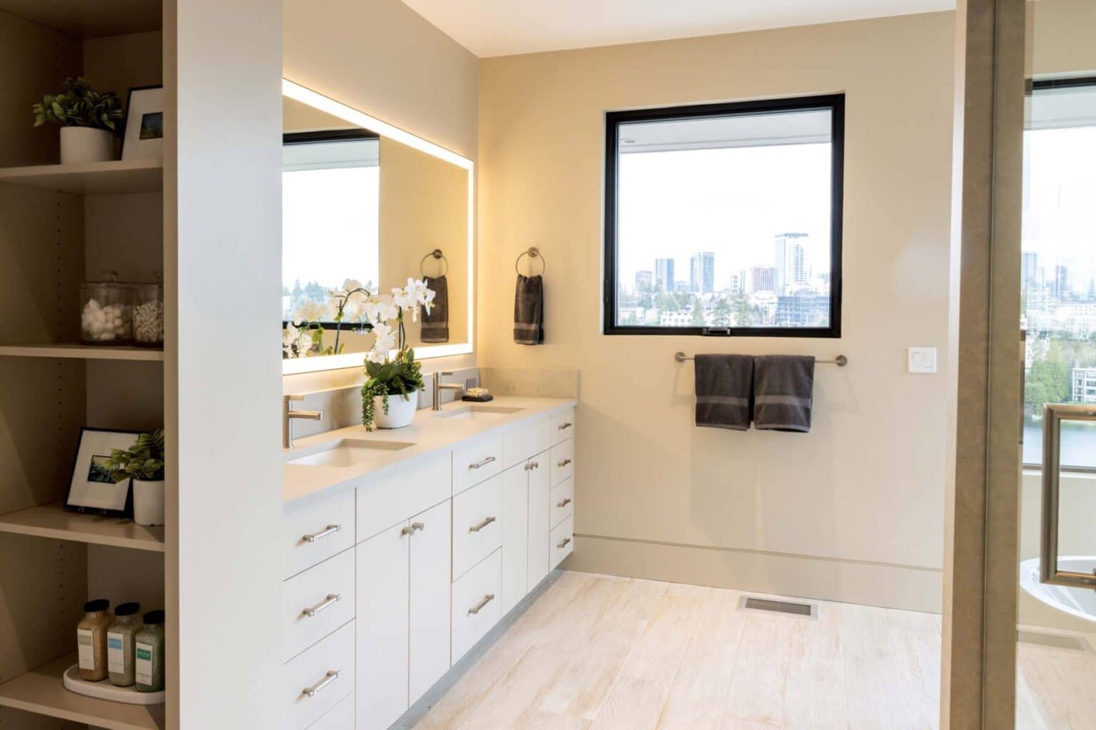 Modern bathroom with a double sink vanity featuring custom finish carpentry Bellevue craftsmanship, large lit mirror, open shelving, two dark towels on a rack, and a window overlooking a city skyline.