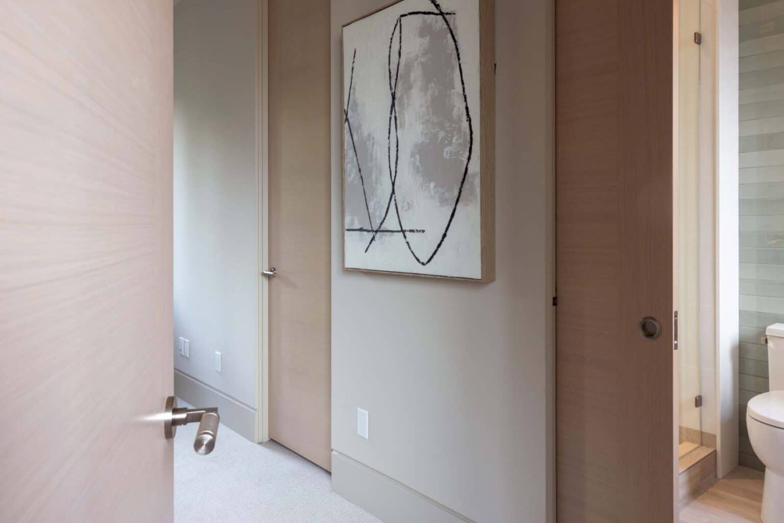 A partially open door reveals a hallway with beige walls, abstract wall art, and a glimpse into a bathroom with striped tiles and a toilet—highlighting the refined touch of custom finish carpentry Bellevue in every detail.