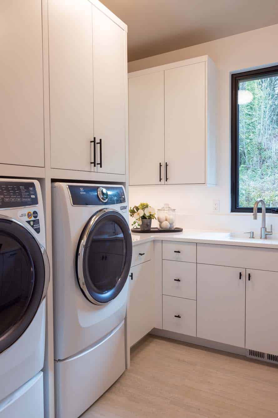 Modern laundry room with stacked washer and dryer, white cabinets, a countertop with decor items, and a window above the sink—showcasing refined custom finish carpentry Bellevue for a polished and stylish look.