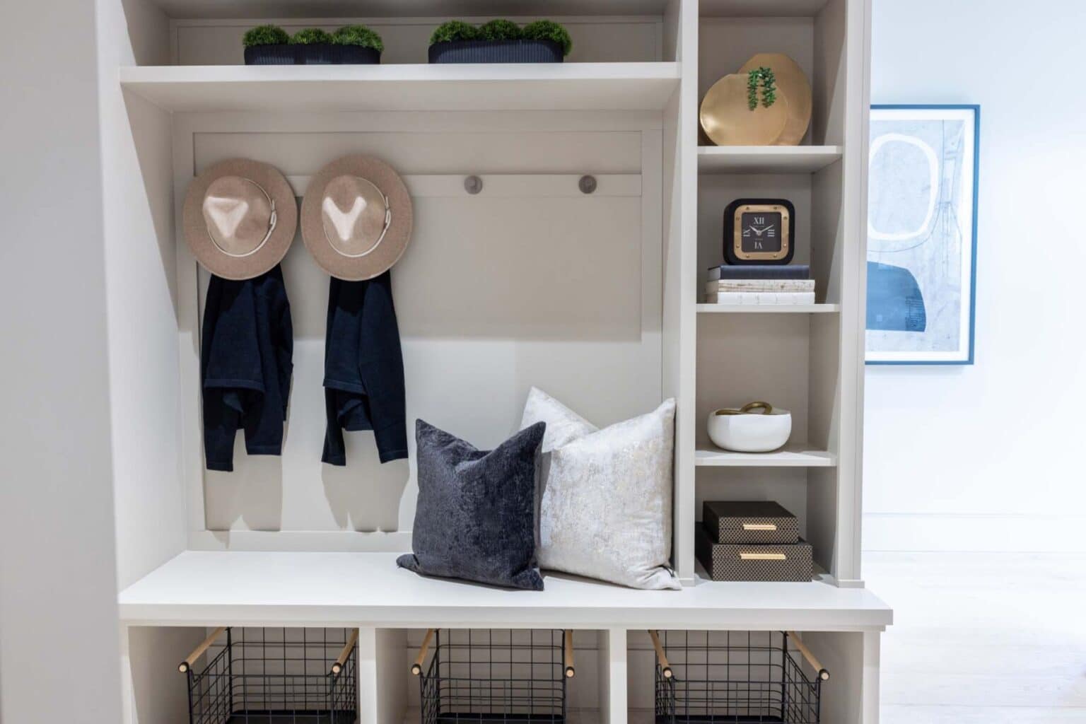 Built-in entryway storage with hooks, hats, coats, pillows on a bench, shelves with decor, wire baskets below, and wall art in the background showcases elegant custom finish carpentry Bellevue homeowners appreciate for both function and style.