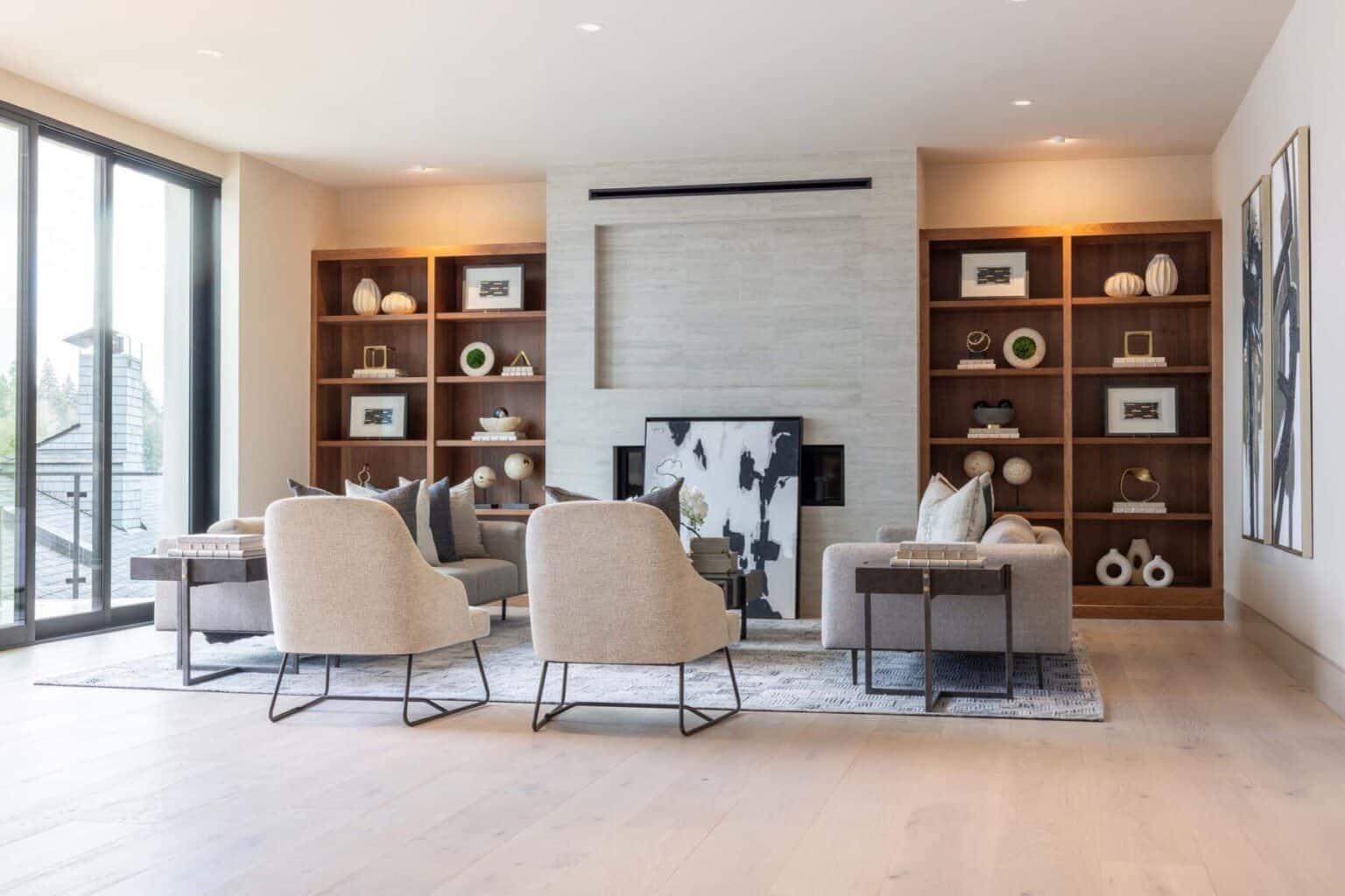 Modern living room with beige armchairs, a fireplace, built-in wooden shelves with decor crafted through custom finish carpentry Bellevue, large windows, and abstract wall art.