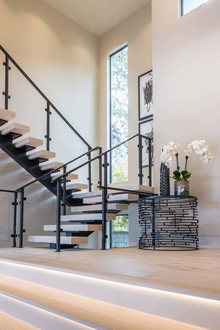 Modern staircase with open wooden steps, black metal railing, and a table with white orchids and decor against a neutral wall with tall windows, showcasing the elegance of custom finish carpentry Bellevue.
