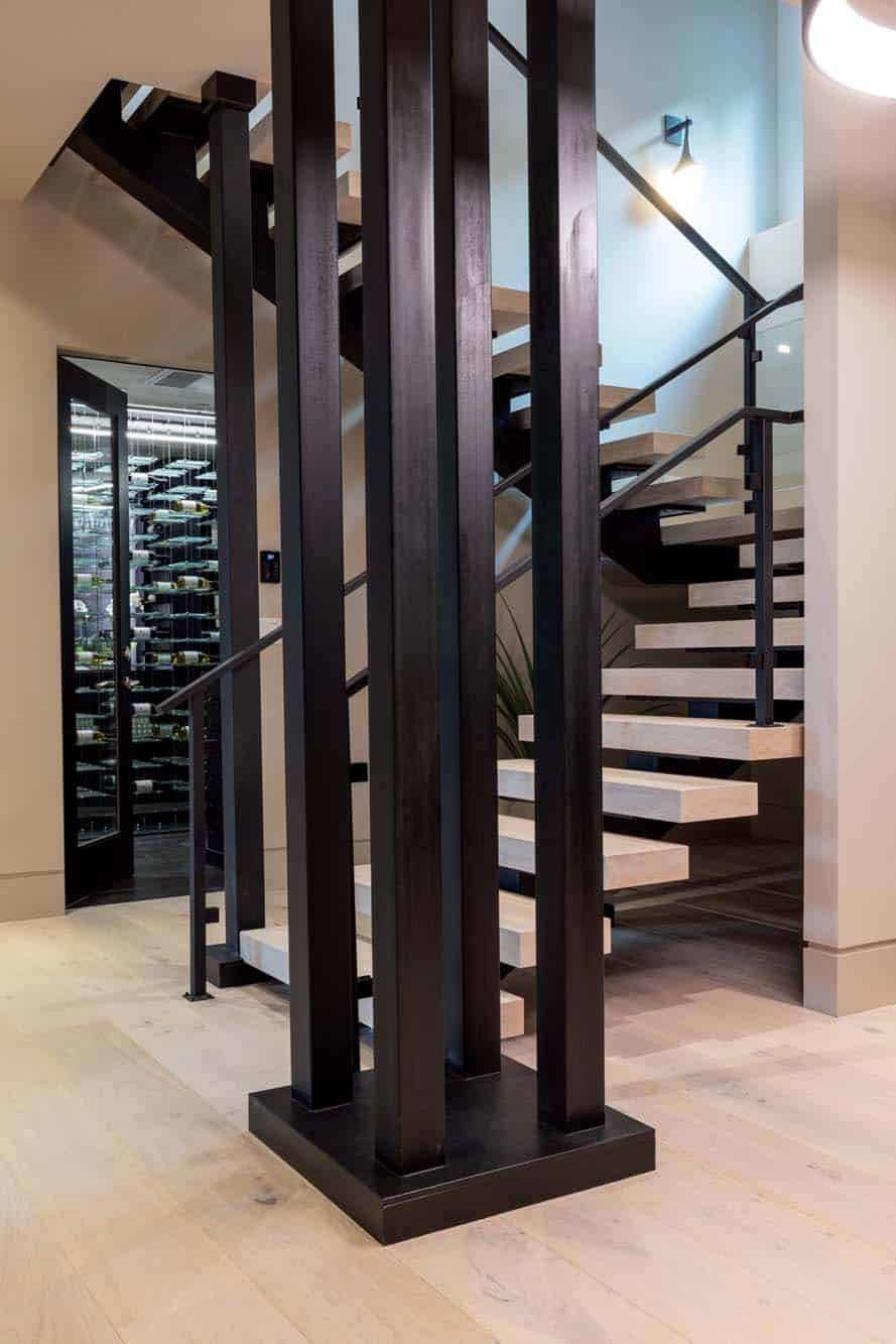 Modern interior featuring floating wood stairs with black metal railings and vertical support beams, showcasing the artistry of custom finish carpentry Bellevue. A glass wine cellar is visible in the background, complemented by light wood flooring throughout.