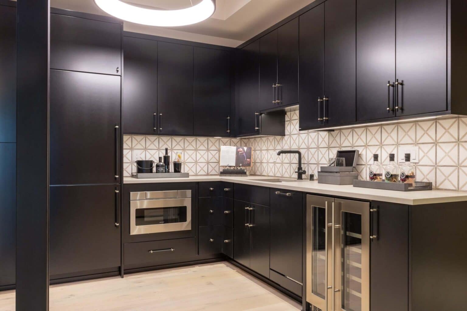 Modern kitchen with black cabinets, geometric tile backsplash, built-in microwave, wine fridge, and countertop appliances; light wood floor and circular ceiling light—all enhanced by custom finish carpentry Bellevue for a refined and cohesive look.