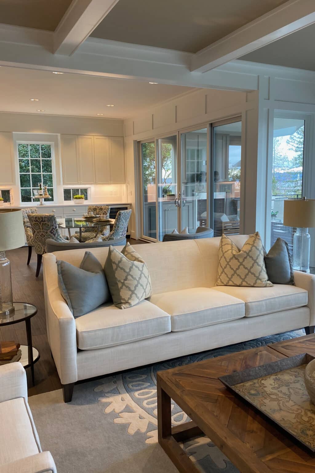 Neutral-toned living room with a beige couch, patterned pillows, wooden coffee table, and an open view into a kitchen and dining area with large windows featuring custom finish carpentry Medina for added elegance.