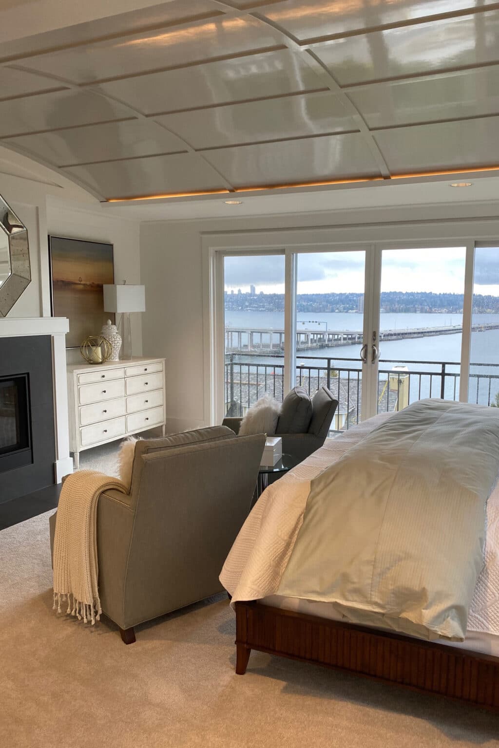 A modern bedroom with a fireplace, armchair, and large bed—enhanced by custom finish carpentry Medina—overlooks a waterfront through sliding glass doors leading to a balcony.