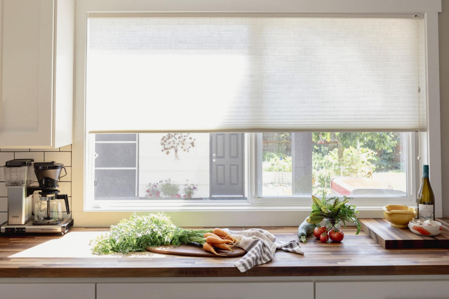 A kitchen countertop with fresh vegetables, a cutting board, a coffee maker, a bottle of wine, and sunlight coming through a window with partially closed blinds highlights the impeccable kitchen finish carpentry West Seattle is known for.