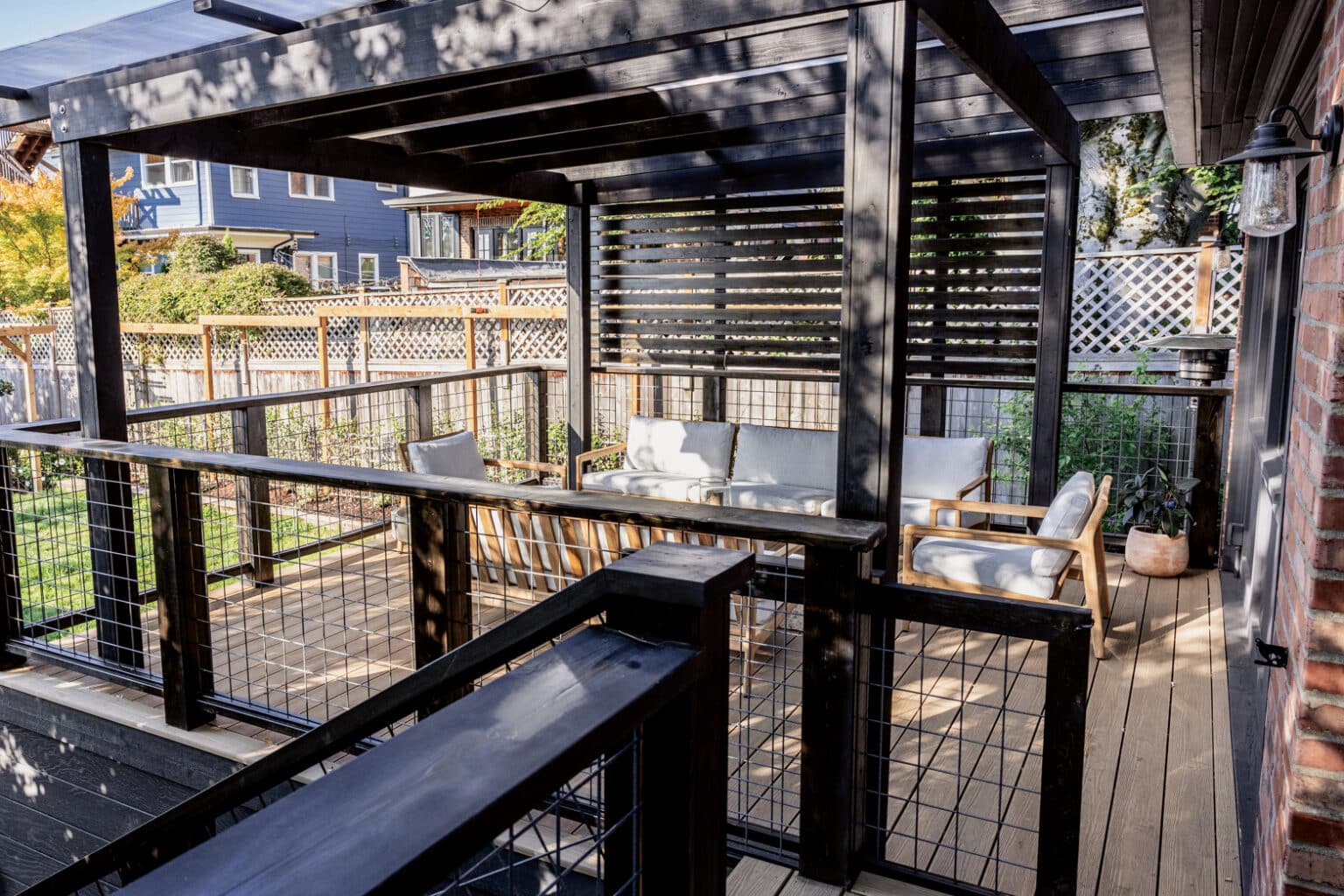 Wood deck with black pergola, modern cable railing, and outdoor seating area with cushions. Expert outdoor finish carpentry in Seattle is showcased in the precise installation of lattice fencing surrounding the deck, which overlooks a beautiful garden.