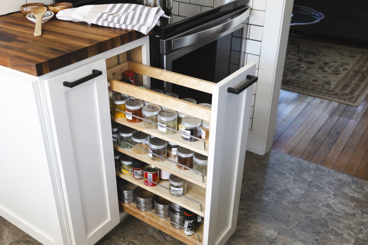 A narrow pull-out kitchen cabinet with three shelves holds jars and tins of spices next to a stove and wooden countertop, showcasing the attention to detail typical of expert kitchen finish carpentry in West Seattle.