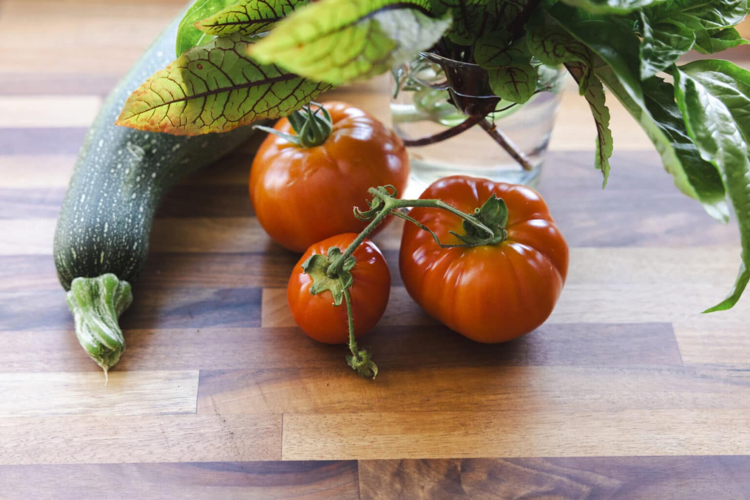 Zucchini, three tomatoes, and a glass vase with leafy greens are arranged on a wooden surface, adding a fresh touch to this kitchen finished with expert carpentry in West Seattle.
