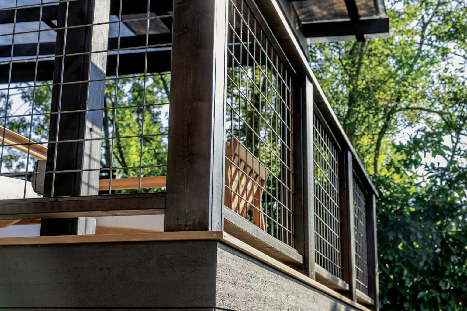Close-up of a modern wooden deck railing with metal grid panels, surrounded by green trees in bright sunlight—showcasing expert outdoor finish carpentry Seattle homeowners appreciate.