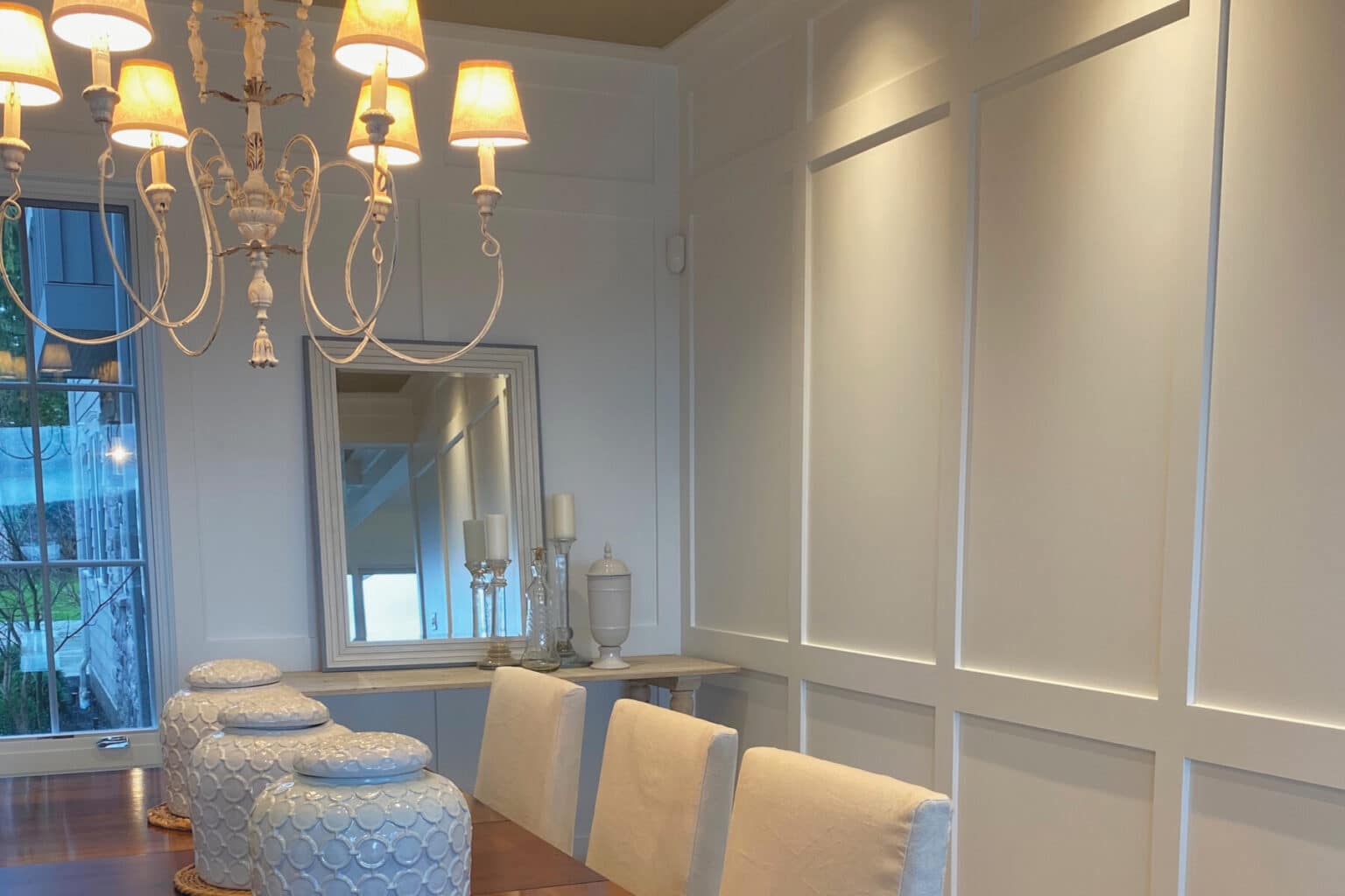 A dining room with a wooden table, four upholstered chairs, decorative jars, a chandelier, a mirror, and candles on a side table showcases the elegance of custom finish carpentry Medina against white paneled walls.