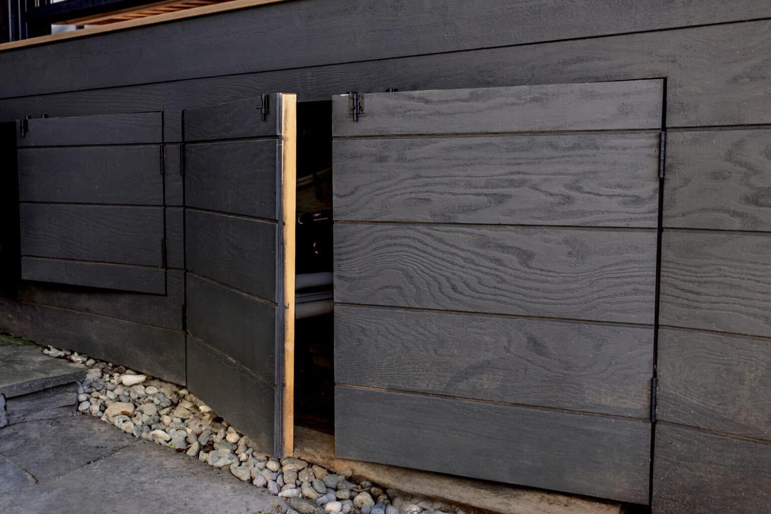 Two dark wooden panels with visible grain, crafted with expert outdoor finish carpentry Seattle is known for, serve as doors or hatches on the side of a building; one is partially open, revealing a small opening beneath the structure. Stones line the ground below.