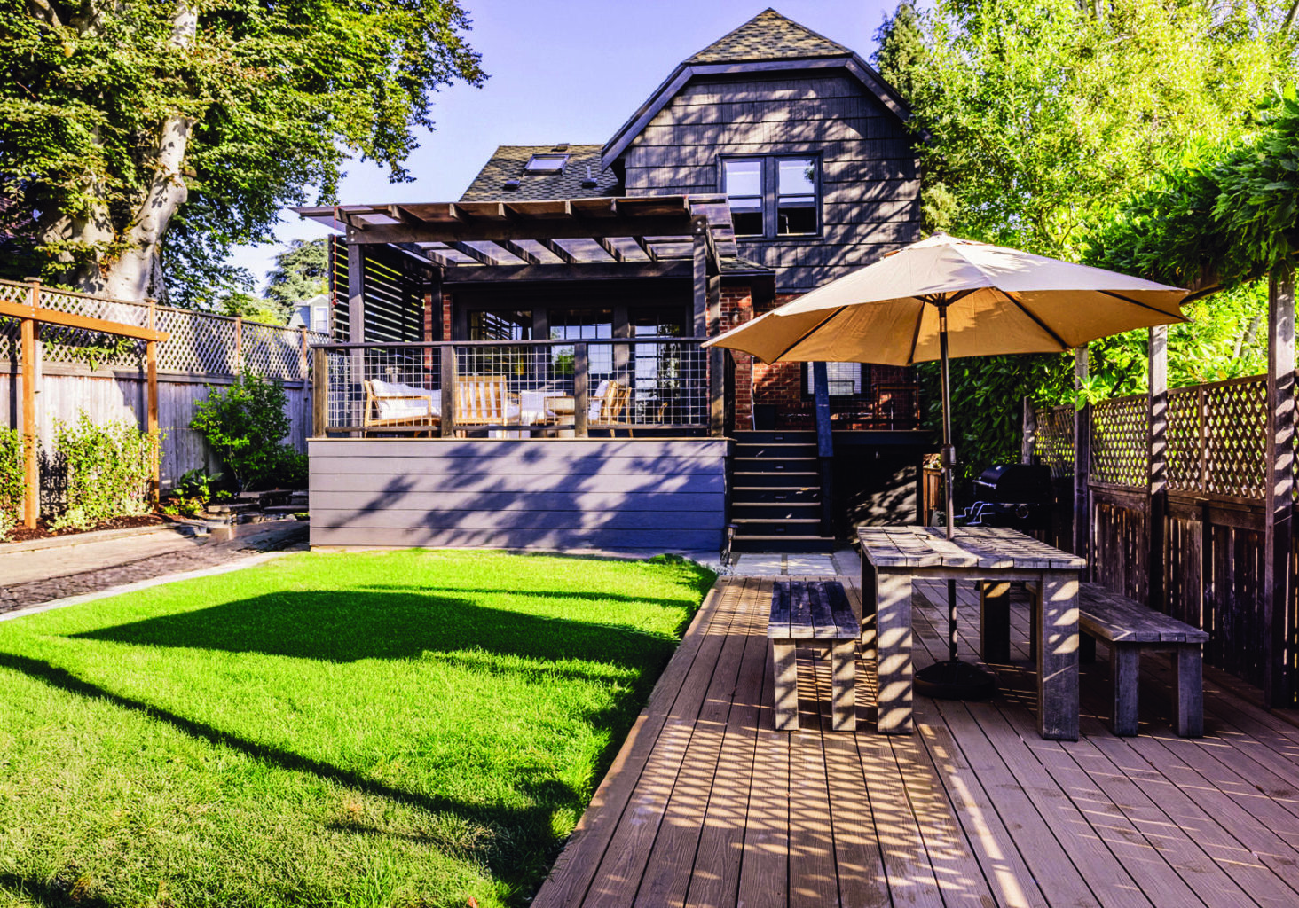 Backyard with green lawn, wooden deck, outdoor dining table with umbrella, and a modern house with a covered patio in the background.