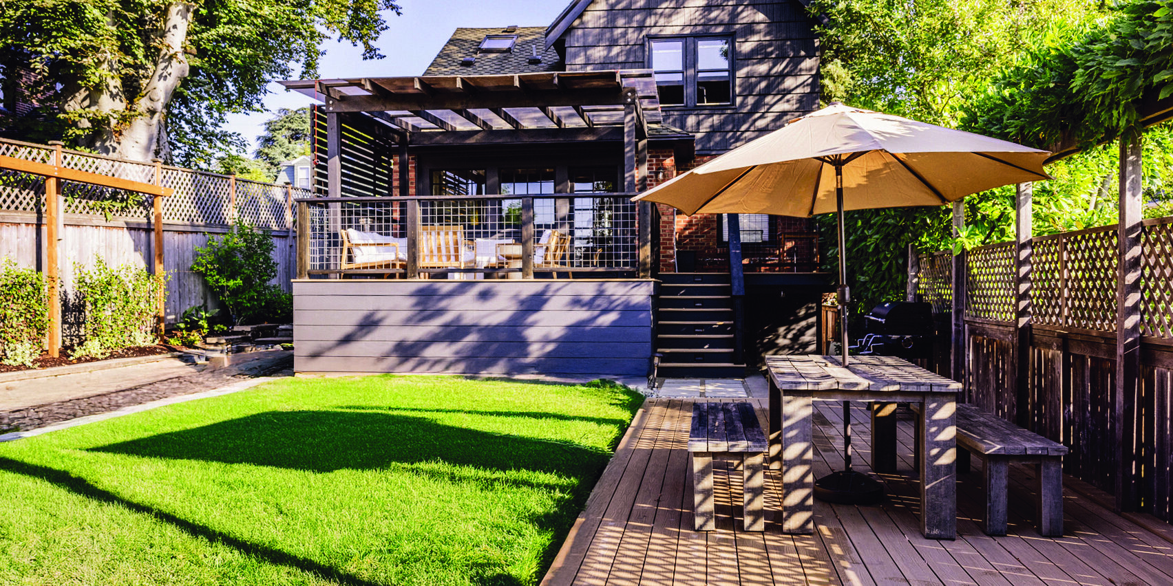 Backyard with green lawn, wooden deck, outdoor dining table with umbrella, and a modern house with a covered patio in the background.