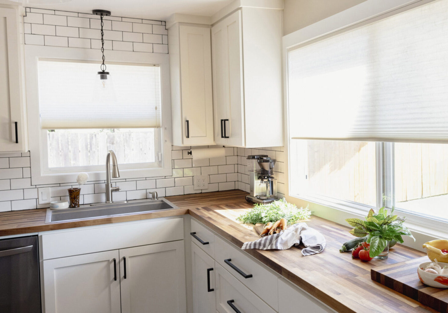Bright kitchen with white cabinets, subway tile backsplash, wood countertops, and fresh vegetables on the counter near a window with blinds.