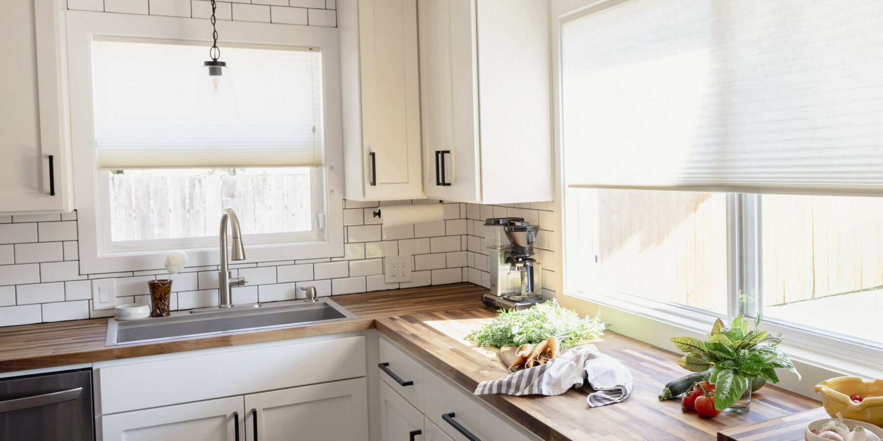 Bright kitchen with white cabinets, subway tile backsplash, wood countertops, and fresh vegetables on the counter near a window with blinds.