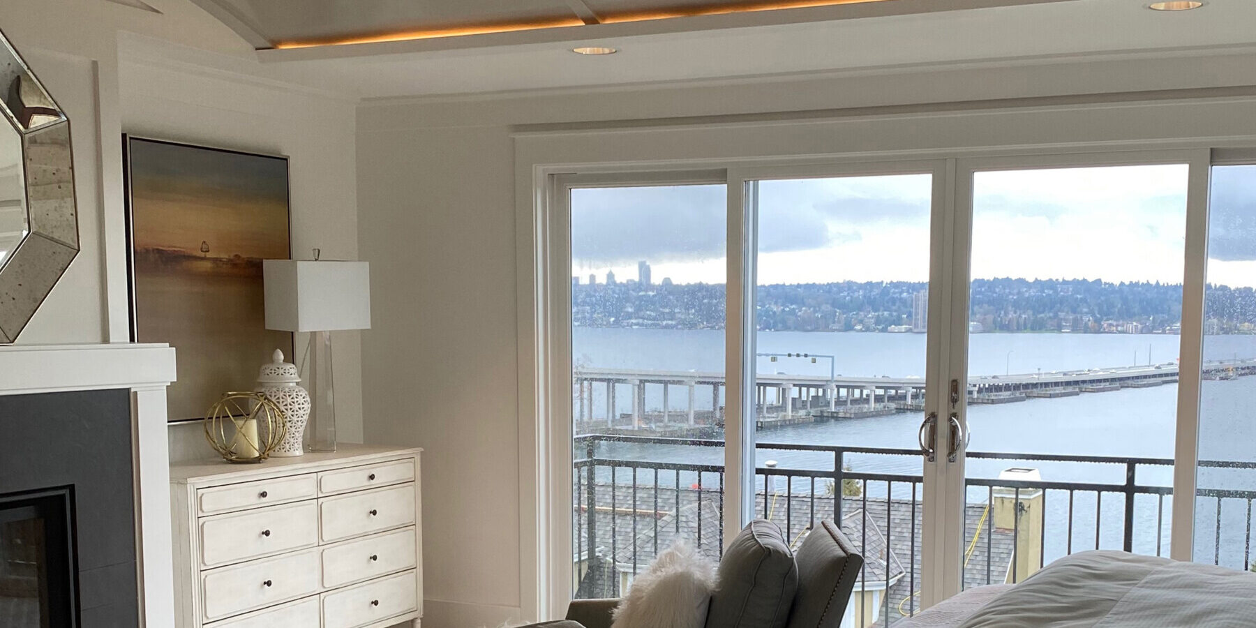 A modern bedroom with light decor features large windows and a balcony overlooking a body of water and a bridge on a cloudy day.